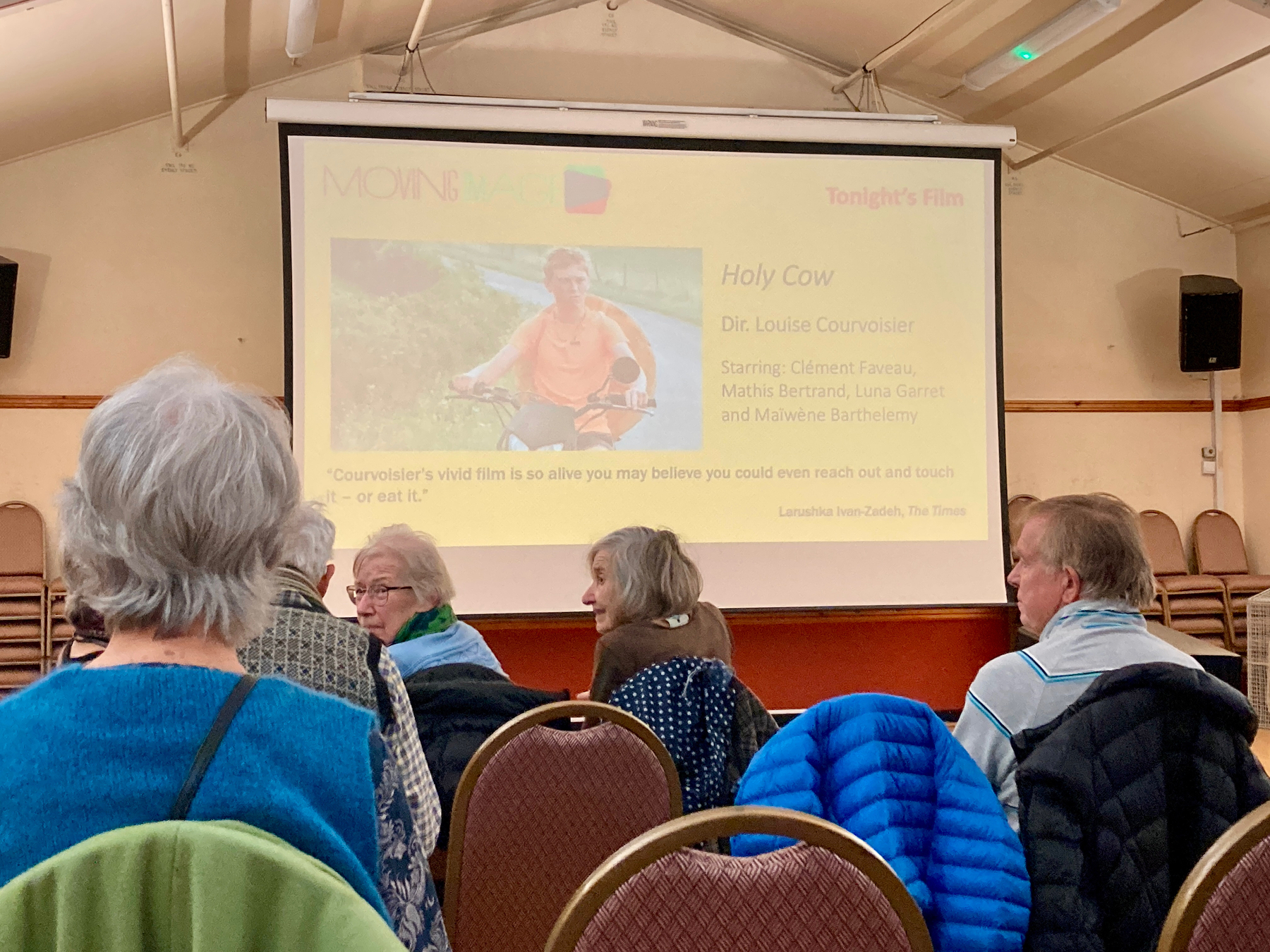 Auto-generated description: People are seated in a room facing a screen displaying information about the film Holy Cow directed by Louise Courvoisier.
